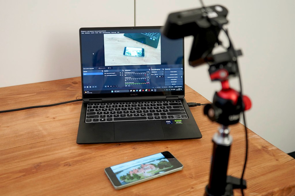 Ein Smartphone liegt vor einem geöffneten Laptop und wird mit einer Kamera gefilmt.