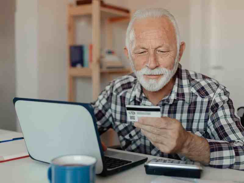 Ein älterer Herr sitzt mit dem Smartphone in der Hand vor einem Laptop.