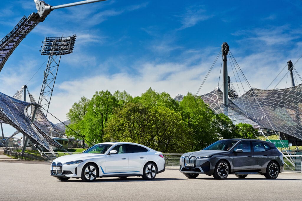 Ein silberner und ein dunkler BMW hintereinander von der Seite auf Platz mit Bäumen und Stadion im Hintergrund