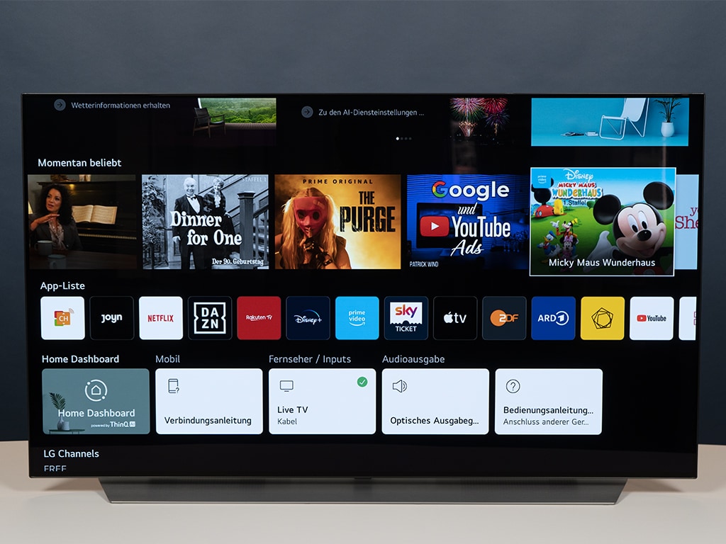 Schwarzer Fernseher von vorne zeigt verschiedene Apps auf weißem Tisch vor grauem Hintergrund