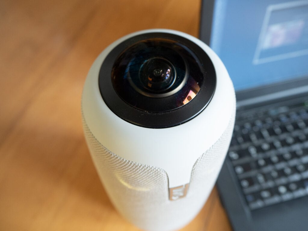 Die zylinderförmige weiße Kamera schräg von oben mit großer schwarzer Kameralinse auf Holztisch neben Notebook