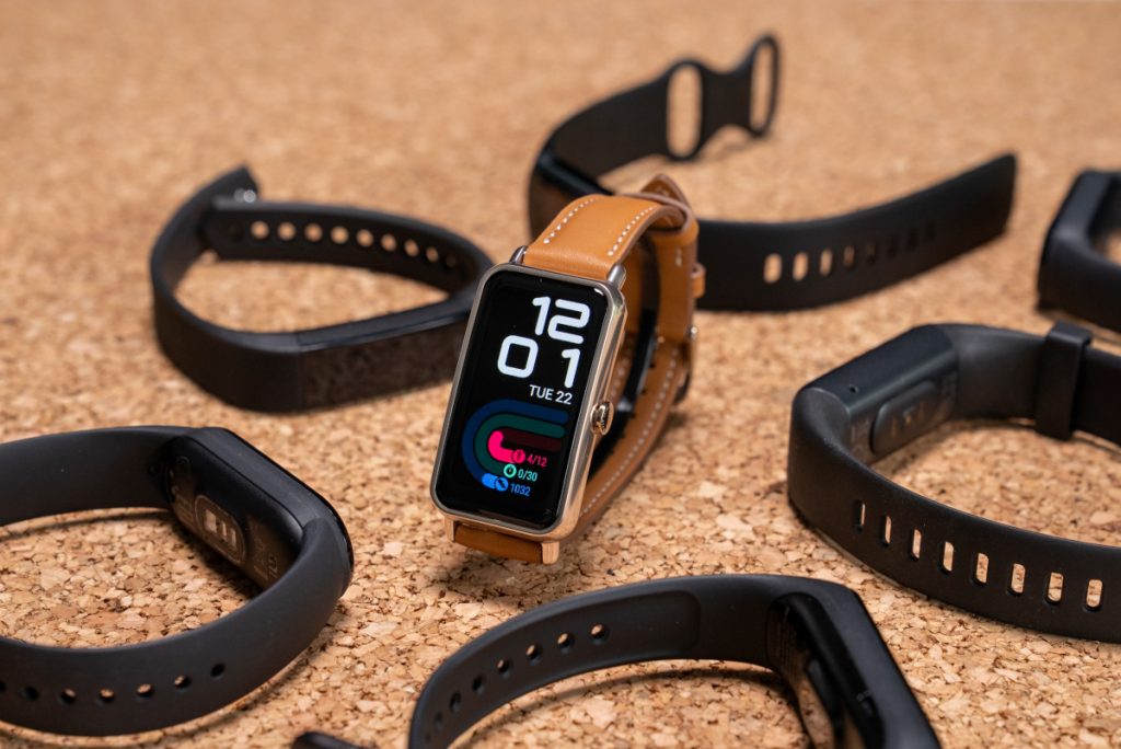 Stehender Fitness-Tracker mit braunem Armband umgeben von liegenden schwarzen Fitness-Trackern