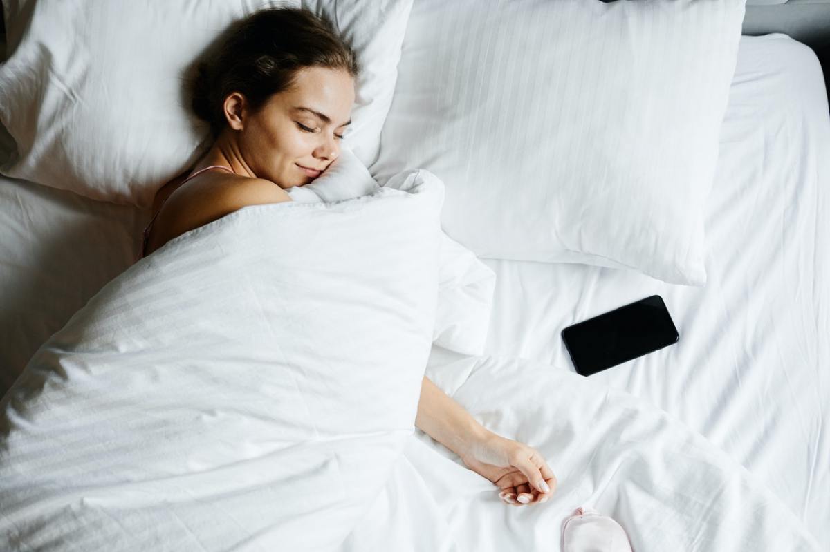 Frau mit weißer Decke zugedeckt im Bett lächelt mit geschlossenen Augen, neben ihr ein Smartphpne
