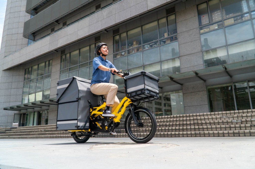 Mann fährt durch städtisches Gebiet auf E-Cargobike, das mit Transportboxen bealden ist.