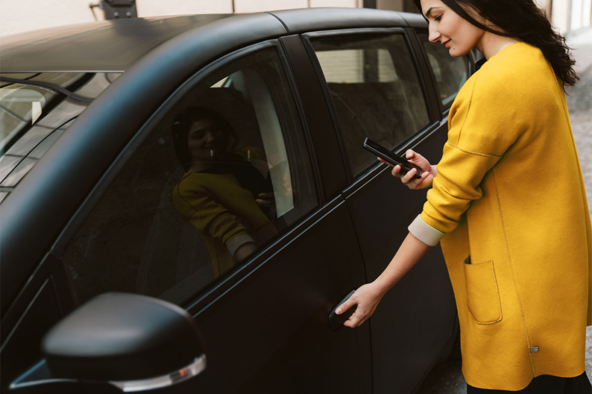 Frau mit Smartphone vor Solarauto. DasSolarauto Sion und normale PKW können über Sono-App miteiender getilet werden.