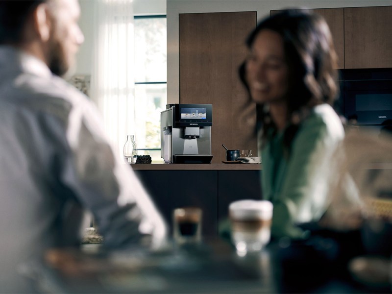 Im Vordergrund sind verschwommen zwei Personen zu sehen, die an einem Tisch sitzen und unterschiedliche Kaffee-Getränke trinken. Im Fokus steht der neue Siemens EQ900-Kaffeevollautomat.