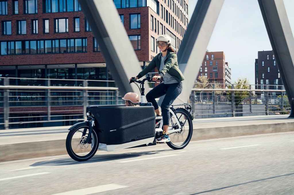 Frau fährt mit E-Cargo-Bike Packstar von riese & Müller über ein Brücke. Im Ladekorb vor ihr sitzt ein Kind.