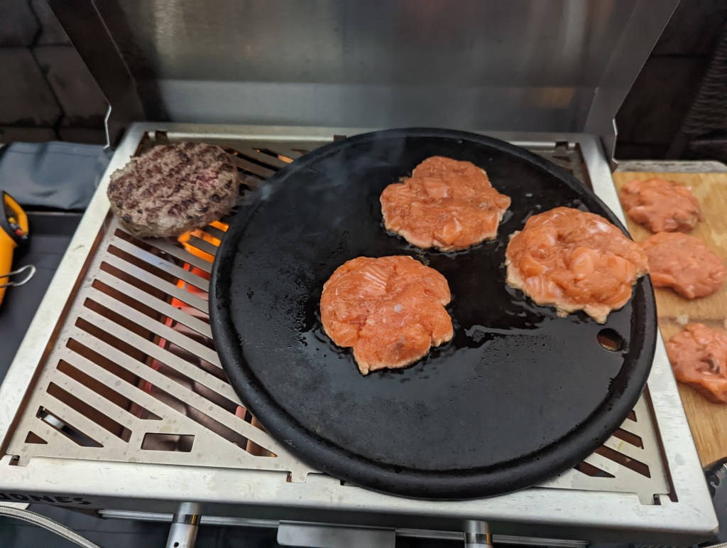 Auf dem Rost des Jones liegen ein Burgerpatty und eine runde Gusseisenplatte mit Lachsfrikadellen