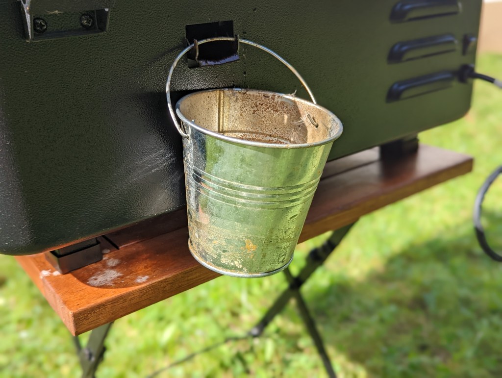 Ein kleiner Blecheimer, der hinten am Grill hängt und das ablaufende Fett auffängt