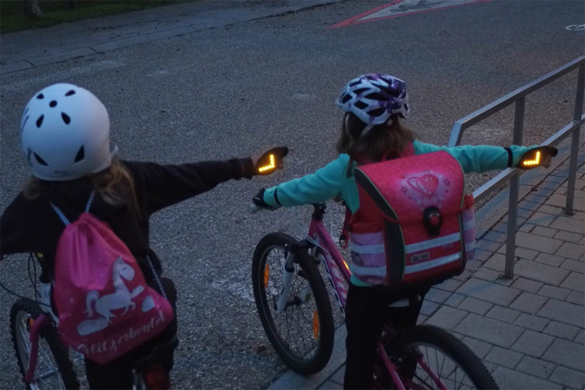 zwei Kinder auf dem Fahrrad, zeigen an, dass sie abbiegen wollen, an der Hand der Überziehhandschuh mit Pfeilblinker