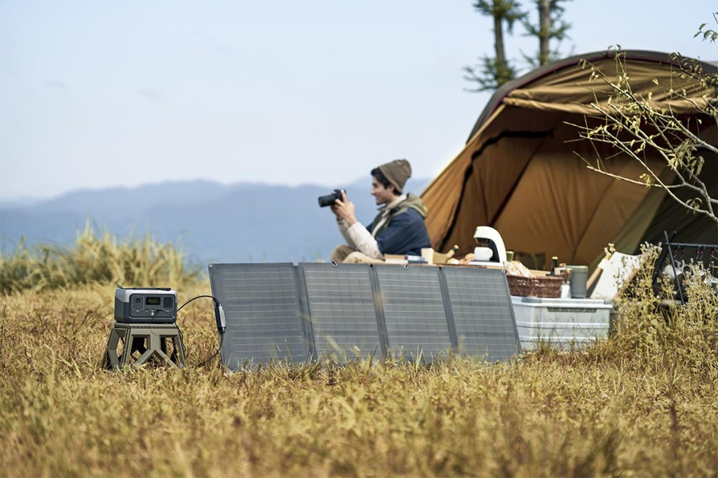 Eine Person sitzt mit Kamera vor einem Zelt. Eine River 2 Power Station wird daneben mit einem Solarpanel geladen.