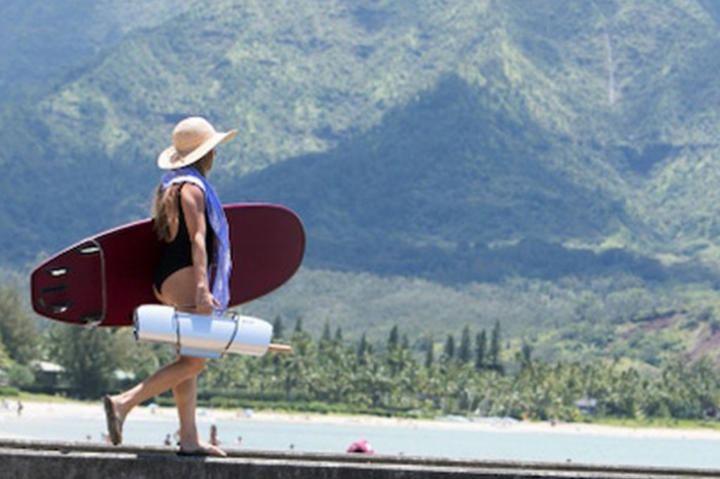 Eine Frau läuft in Bademontur auf einem Steg auf einem See mit Bergen im Hintergrund. Sie trägt ein Surfbrett und den GoSun Solargrill.