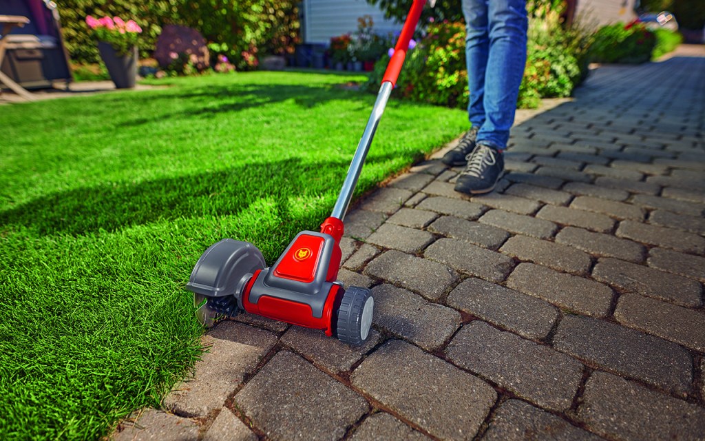 Detailaufnahme eines manuellen Rasen-Kantenschneider im Einsatz. Das Gartenwerkzeug wird mit einer Verlängerung über einen gepflasterten Weg geschoben und schneidet dabei mit der Außenseite eine Rasenkante.