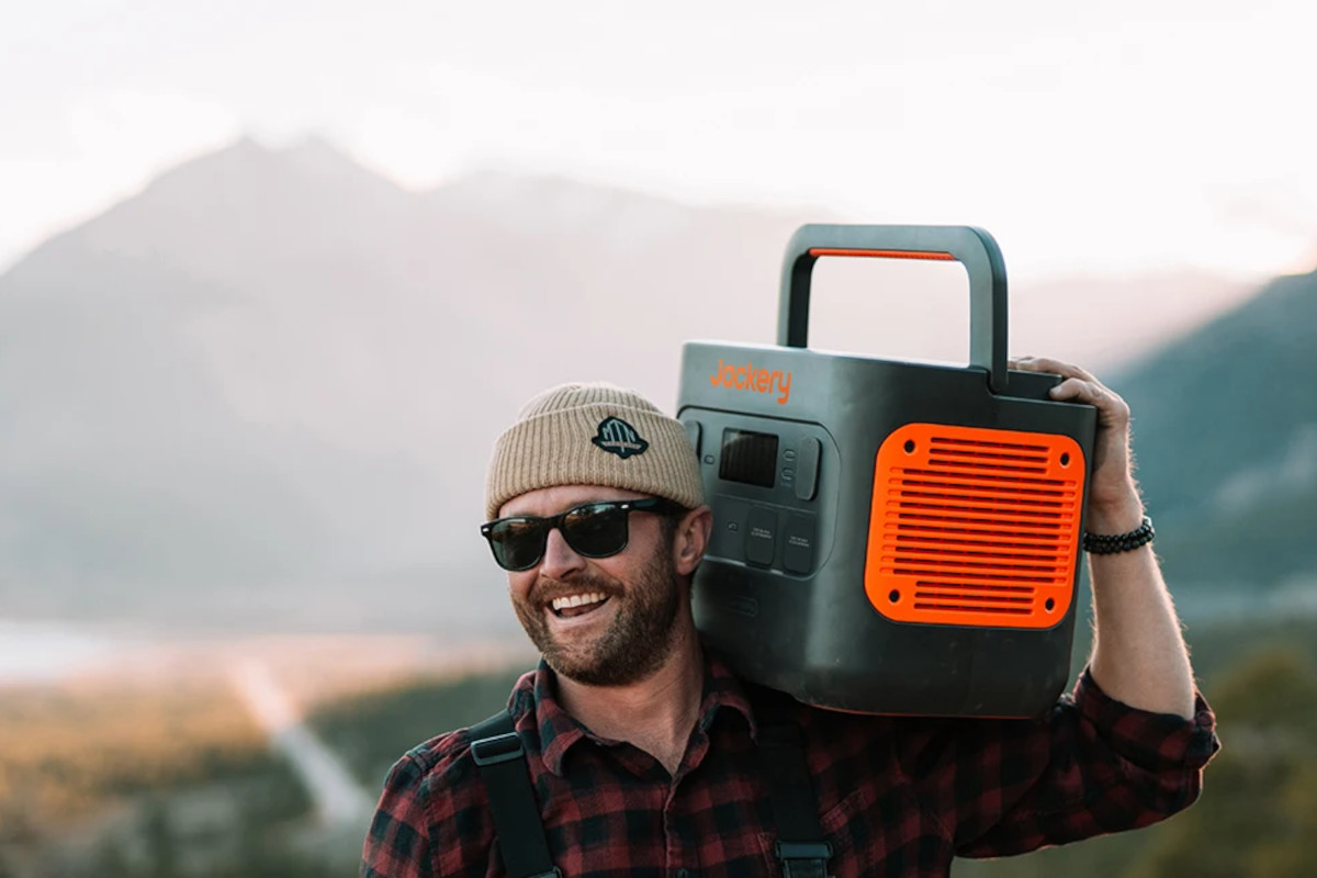 Mann mit Sonnenbrille und Mütze trägt Jackery Power Station in schwarz orange auf Schulter vor Bergkulisse