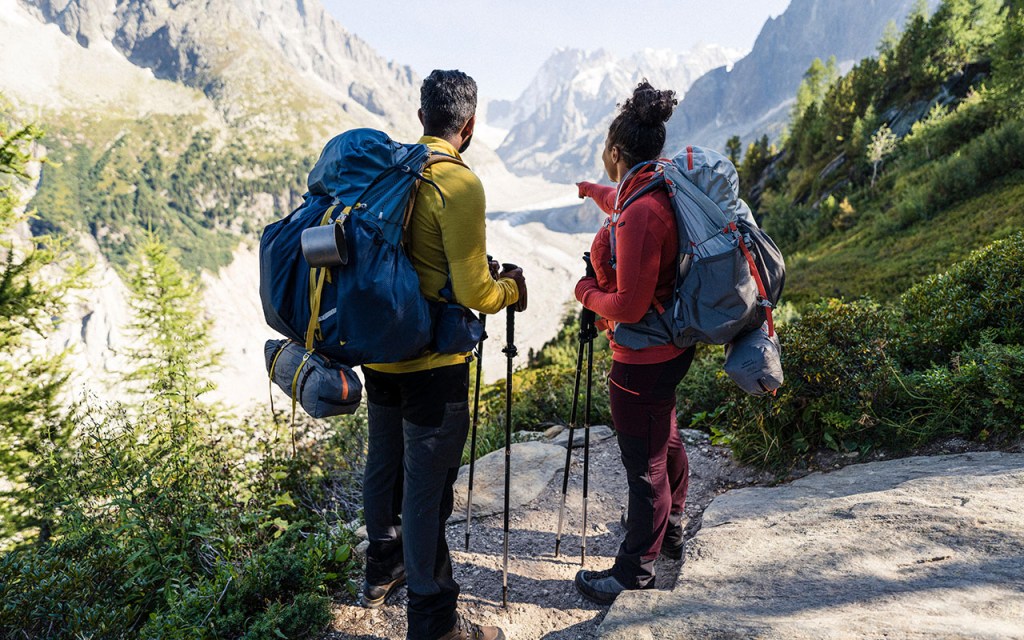 Frau und Mann stehen mit Wanderausrüstung auf einem Weg vor Gebirgslandschaft.