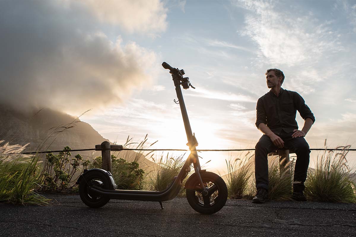 Mann sitzt auf einer Stange in der Natur bei aufgehender Sonne, vor ihm sei E-Scooter