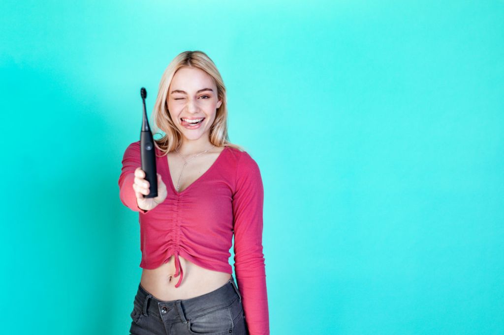 Eine blonde Frau vor türkisem Hintergrund hält mit verschmitztem Gesichtsausdruck eine happybrush ECO Vibe 3 in die Kamera.