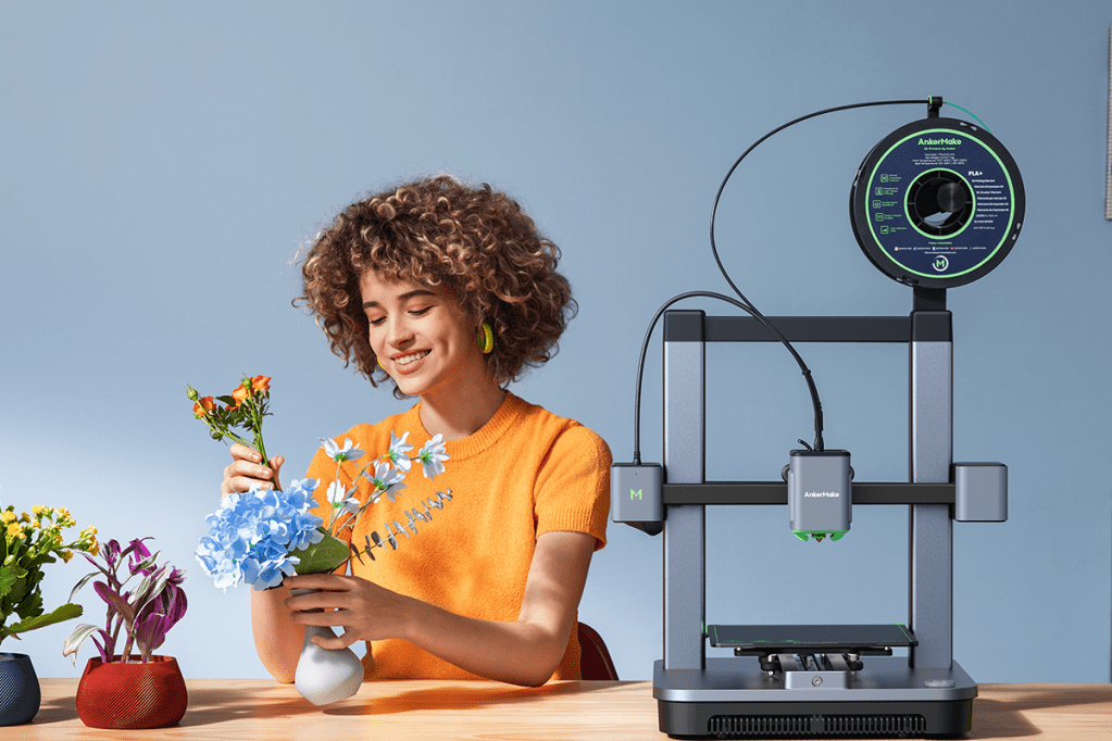 Eine Frau neben dem 3D-Drucker AnkerMake m5c in der der Hand eine gedruckte Vase mit einer Blume.