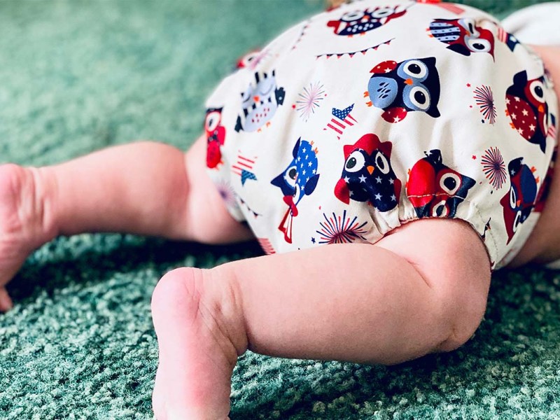 Nahaufnahme eines Babys, das man bauchabwärts auf dem Bauch liegend sieht, es trägt eine Windel mit Eulenmuster