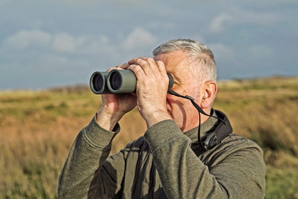 Man mit Fernglas steht in grüner Jacke in der Natur herum.