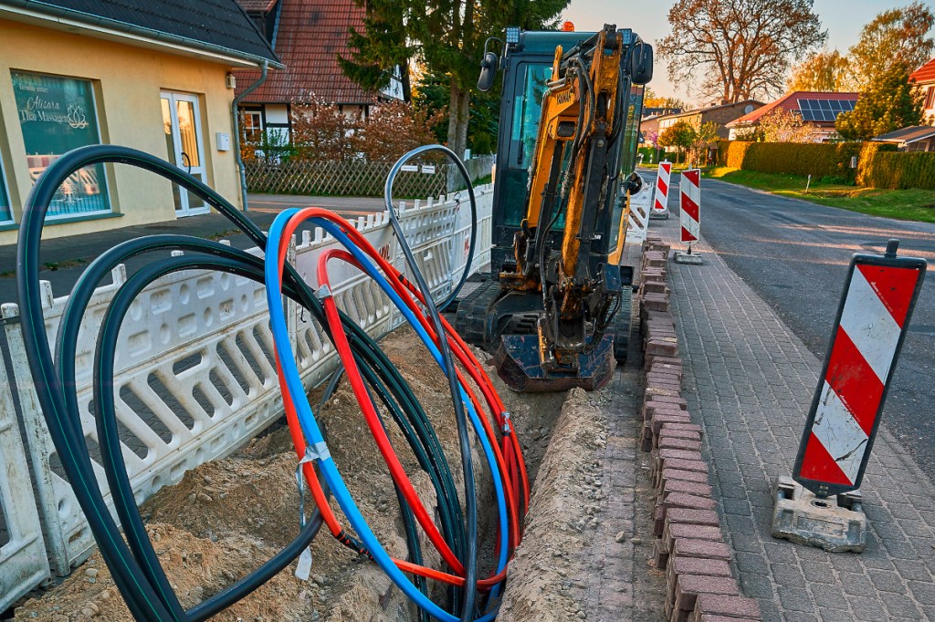 Baustelle Gehweg offen mit verschiedenen großen Kabeln eingerollt, die herausragen, im Hintergrund Bagger und Haus