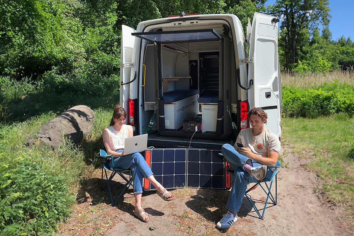 Zwei Personen sitzen vor einem Transporter, der mit einem PlugVan-Modul ausgestattet ist. Außerdem nutzen sie mobile Endgeräte, die mit einer Jackery Powerstation versorgt werden.