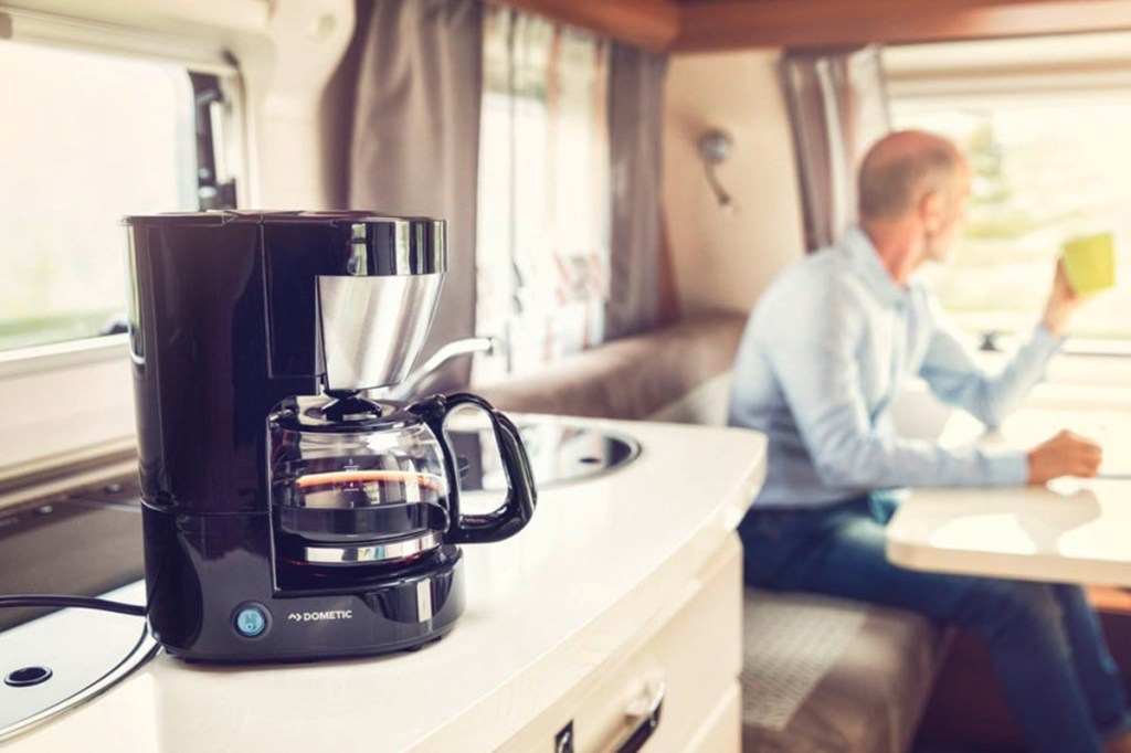 Kaffeemaschine in einem Wohnwagen, dahinter sitzt ein Mann, der einen Kaffee trinkt. 