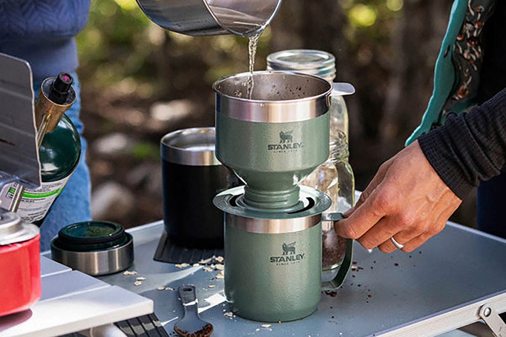 Kaffee wird im Freien auf einem Campingplatz aufgebrüht, indem heißes Wasser in einen Stahlfilter gegossen wird, der auf einem Thermosbecher steht.