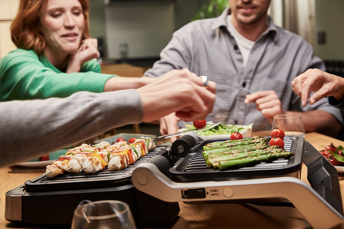 Mehrere Personen sitzen an einem Esstisch, auf dem ein Tefal OptiGrill 4in1 Gemüse und Fleischspieße zubereitet.