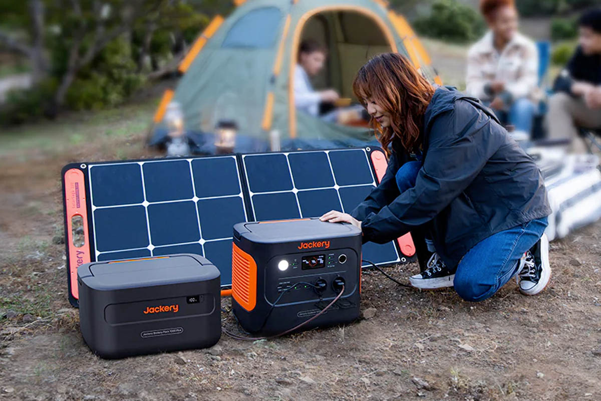 Eine Person schließt ein Solarpanel an die Jackery Explorer 1000 Plus an, die bereits mit einem Zusatz-Akku erweitert ist. Im Hintergrund ist eine Camping-Szene.
