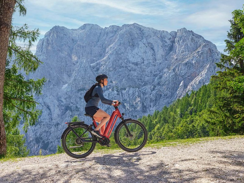 Frau fährt mit einem roten E-Bike auf einer Straße, im Hintergrund Bergkulisse im Sonnenschein