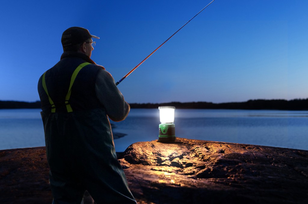 Mann schräg von hinten im Dunkeln hält ausgeworfene Angel in See, vor ihm ein Felsen auf dem eine eingeschaltete Camping-Laterne leuchtet
