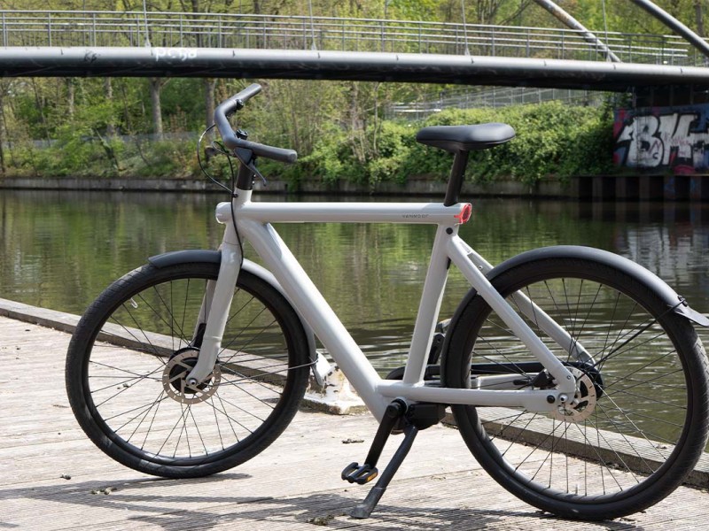 weißes E-Bike auf einem Steg stehend, ein Fluss und eine Brücke im Hintergrund