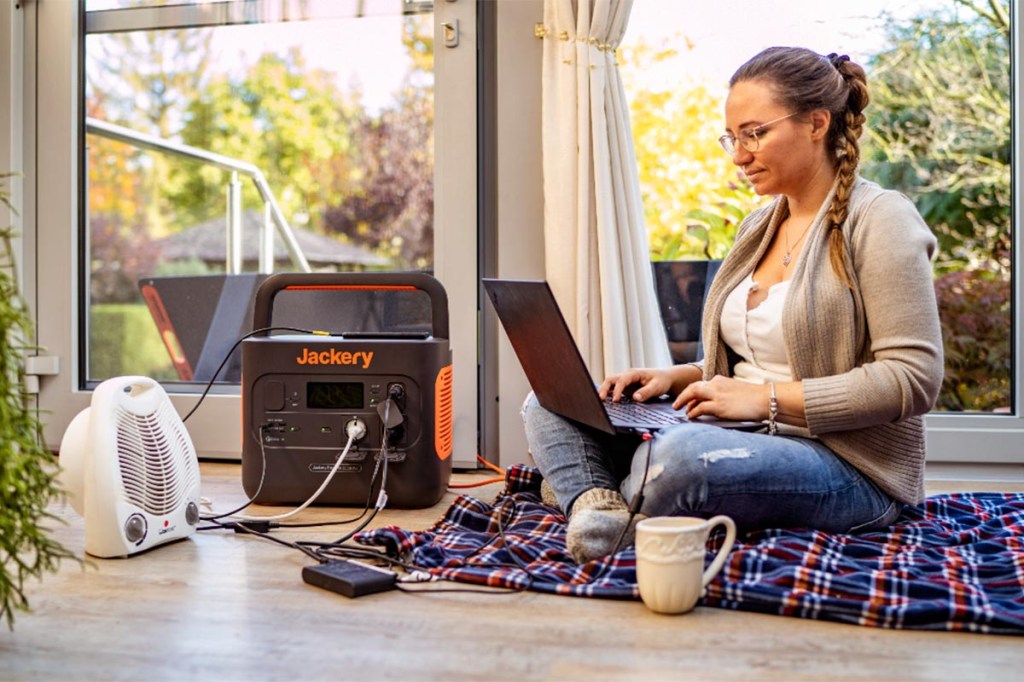 Eine Frau arbeitet auf dem Boden am Laptop und betreibt mit einer Jackery Powerstation einen Heizlüfter und ihren Laptop.