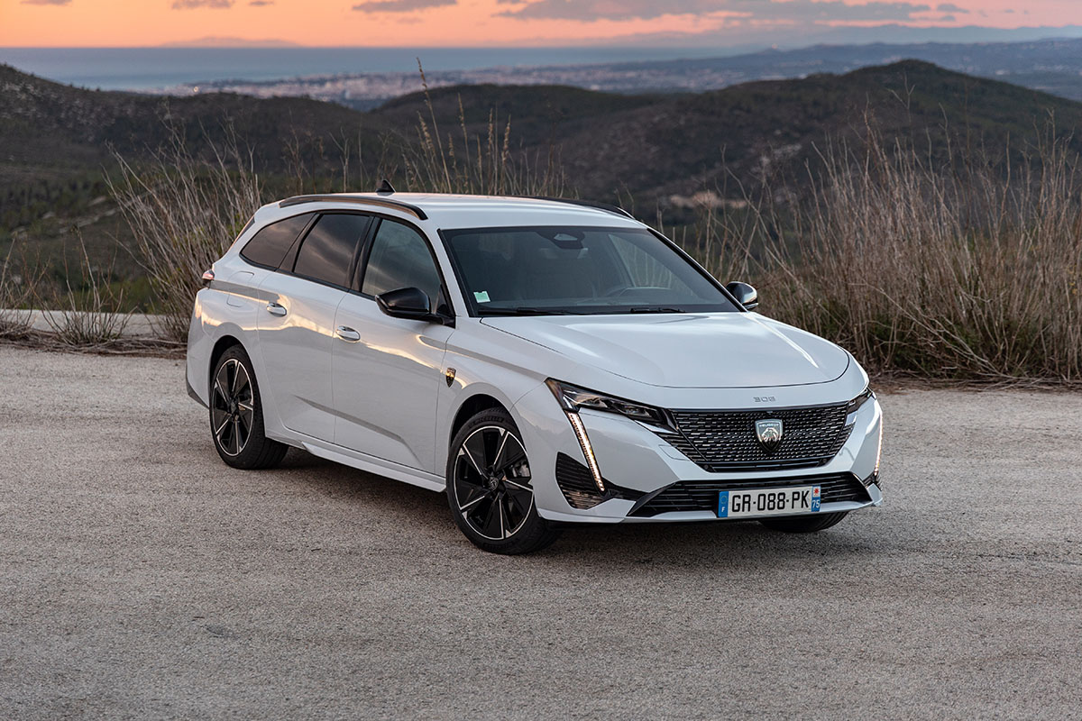 E-Auto Kombi Peugeot E-308 SW in schräger Ansicht von vorn auf einem Parkplatz vor bergiger Kulisse während eines Sonnenuntergangs.