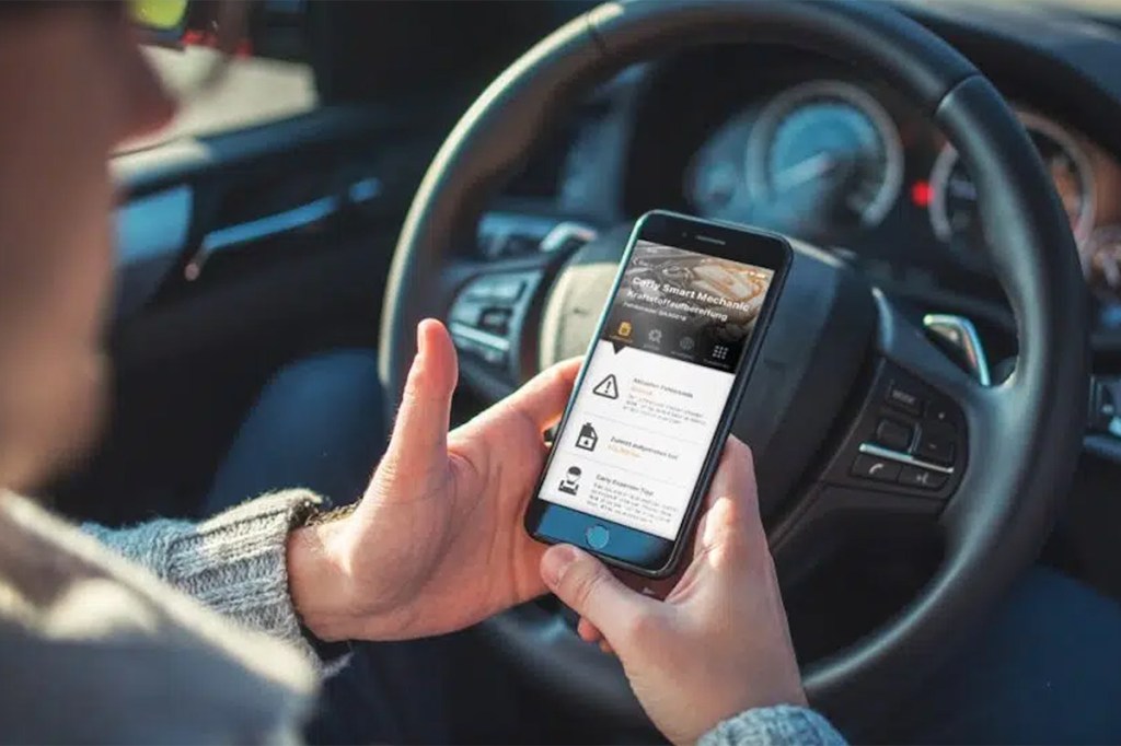 Foto einer Person, die ein Smartphone in der Hand hat und dabei hinterm Steuer sitzt.