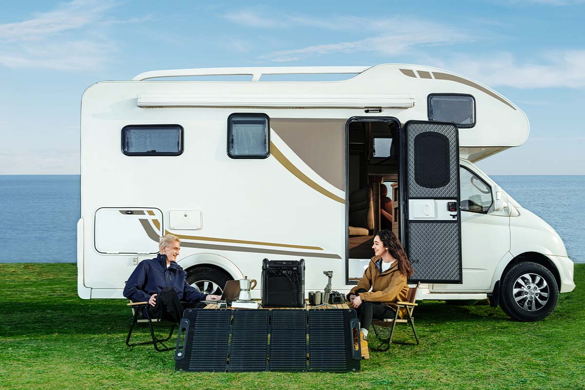 Campervan auf einer Wiese stehend, davor sitzen zwei Personen an einem Tisch, vor ihnen steht ein Solarpanel und eine Powerstation