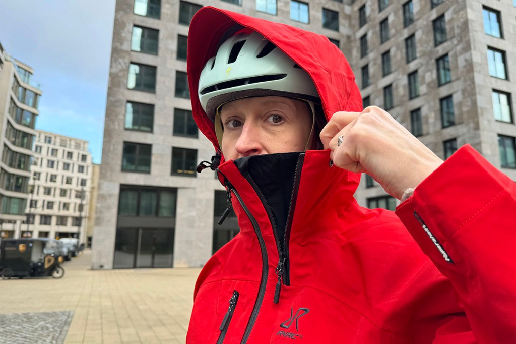 Frau Mit Regenjacke und Fahrradhelm, über dem die Kapuze der Jacke gezogen ist, blickt den Betrachter des Bildes an.