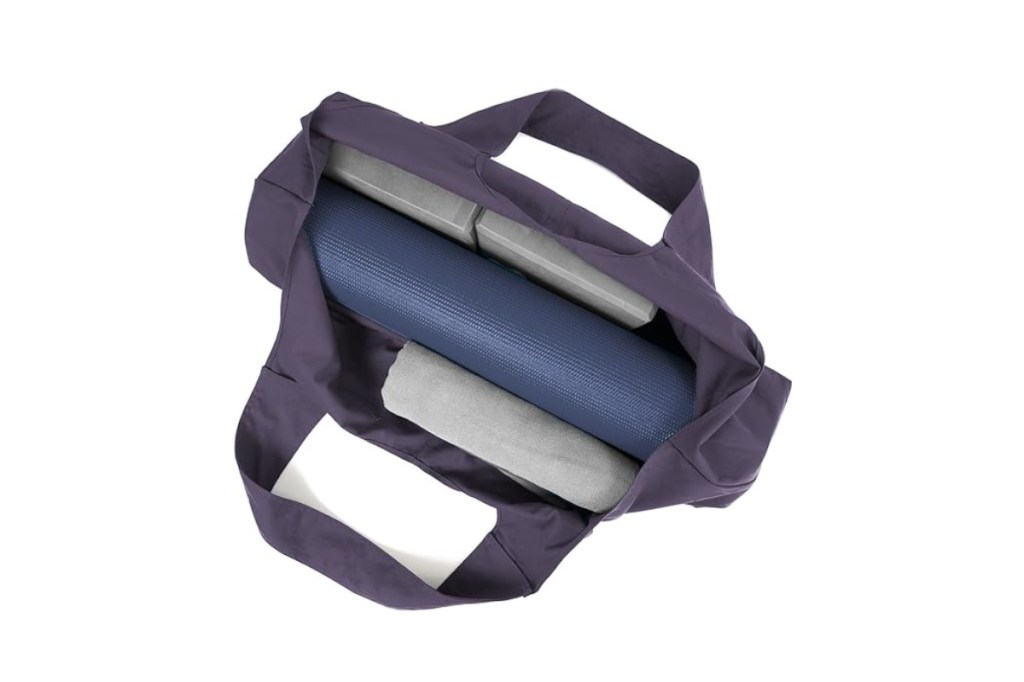 Lila geöffnete Stofftasche länglich von oben mit blauer Yogamatte drin und rechts und links graue Blöcke und Kissen