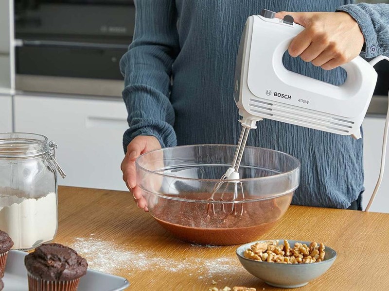 Person im Anschnitt, hat einen Handmixer in der Hand und rührt einen braunen Teig