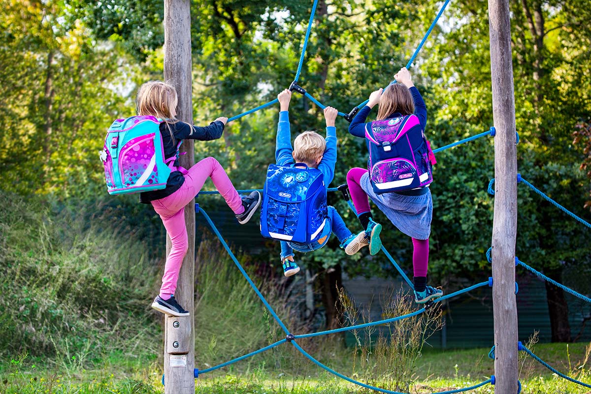 Drei Kinder von hinten, die mit Ranzen auf dem Rücken ein Klettergerüst hochklettern