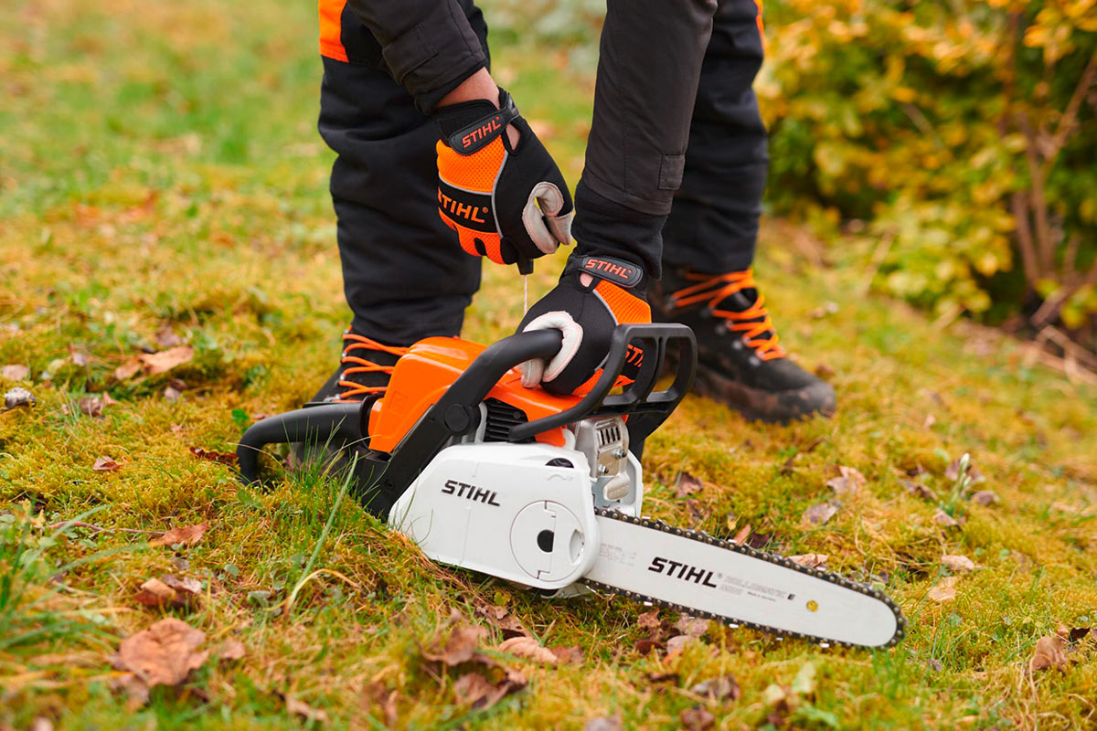 Benzin-Kettensäge von Stihl, auf einer Wiese, dahinter ein Menschen in schwarz-oranger Sicherheitskleidung, der versucht, die Kettensäge zu starten.