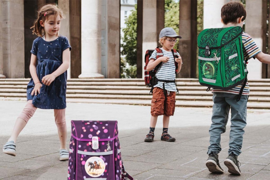 Drei Kinder spielen auf einem Hof, zwei haben einen Ranzen auf dem rücken, ein dritter Ranzen steht auf dem Boden