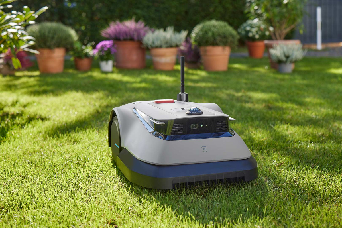 Ecovacs Goat G1-800 Mähroboter ohne Begrenzungskabel in Grau schwarz schräg von vorne auf grünem Rasen, im Hintergrund Blumenkübel mit grünen und lila Blumen