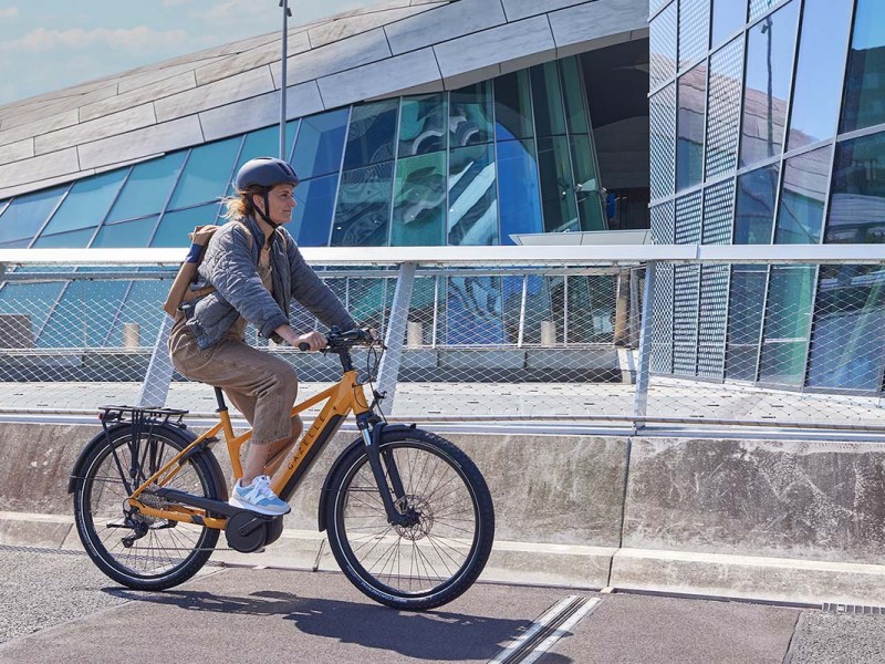 Frau fährt mit einem gelben E-Bike an einer Häuserfront entlang