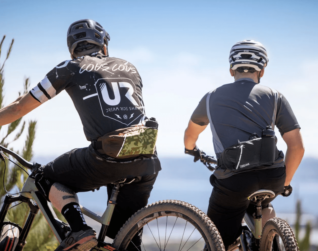 Blick auf die Rücken zwei Mountainbiker auf ihren Fahrrädern welche den CAMELBAK Trinkgürtel umgeschnallt haben.