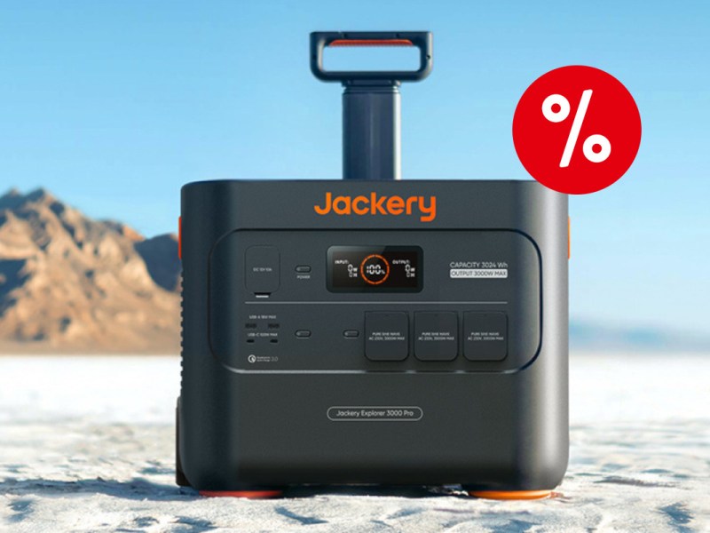 Schwarz orange Jackery Powerstation von vorne mit ausgefahrenem Griff auf weißem Sand vor blauem Himmel und Felsen im Hintergrund mit rotem Prozentbutton oben rechts