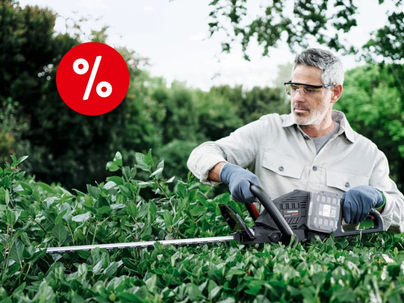 Grauhaariger Mann in weißem Shirt mit grauen Schutzhandschuhen und durchsichtiger SChutzbrille steht hinter grüner Hecke und hält eine graue Parkside-Heckenschere an die Äste, im Hintergrund weitere Hecken und Bäume mit rotem Prozentbutton oben links