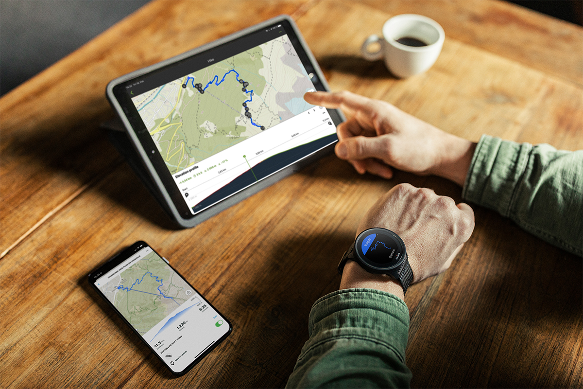 Overshoulder-Blick: Finger zeigt auf ein Tablet, daneben liegt ein Smartphone, zudem sieht man am linken Arm der Person eine Smartwatch. Alle Bildschirme zeigen eine Routenplanung