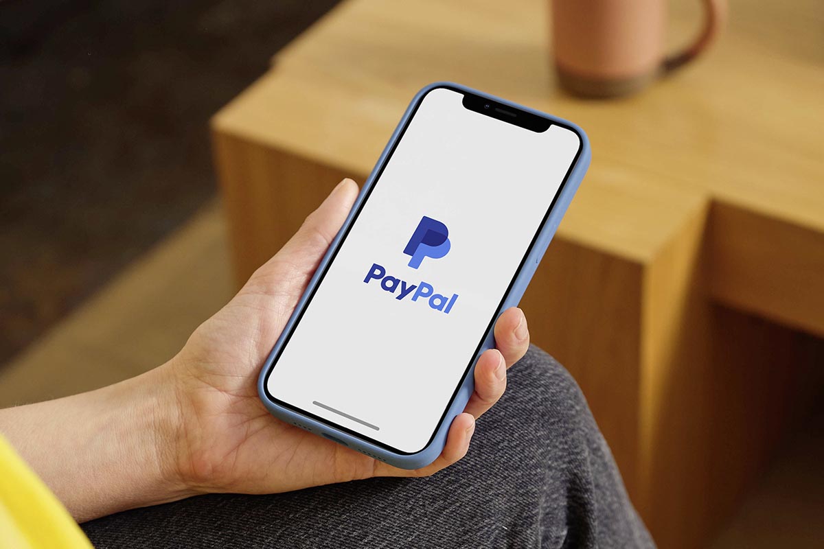 Eine Person hält ein Smartphone in der Hand, auf dem das Paypal Logo zu sehen ist.